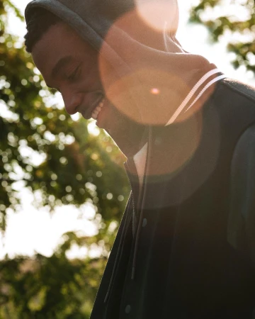 Close-up portrait of a Black man smiling outdoors wearing a dark hooded jacket in bright sunlight.