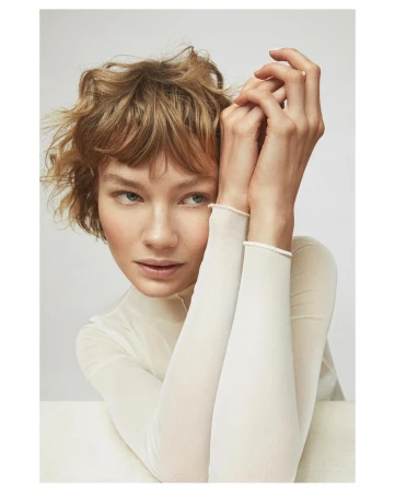 Close-up portrait of a woman with short curly brown hair wearing a white sheer top, holding her hands together near her face against a white background.