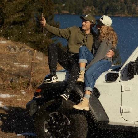 A couple poses atop a white Jeep against a picturesque backdrop of a lake and trees, capturing an outdoorsy, candid moment together.