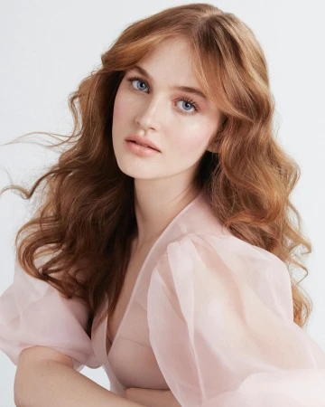Portrait of a woman with long wavy reddish-blonde hair wearing a sheer pale pink blouse against a white background