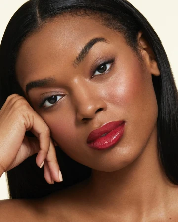 Close-up beauty portrait of a Black woman with glossy red lips and dark hair resting hand near cheek against a neutral light background.