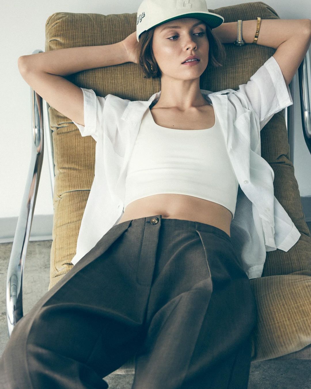 A young woman with short brown hair poses in a white crop top, white button-down shirt, and brown pants while reclining in a chair.