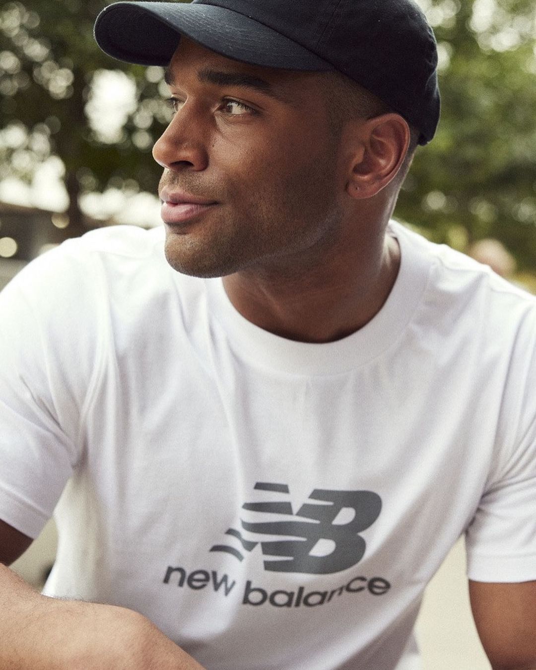 Portrait of a dark-skinned adult male wearing a black baseball cap and a white New Balance branded t-shirt, gazing to his left.