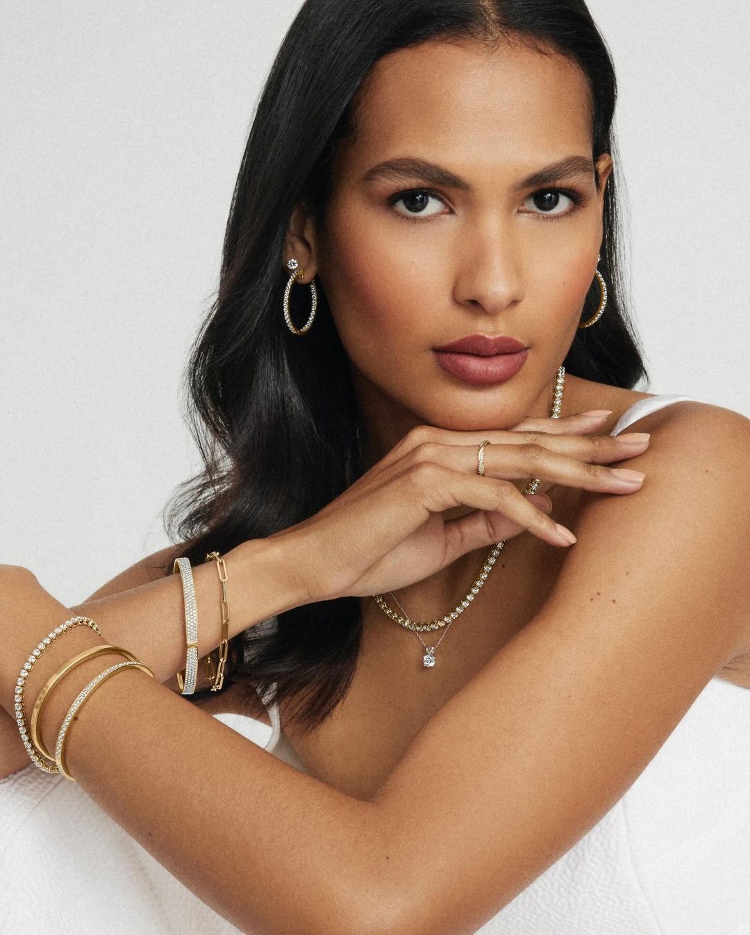 Portrait of a brunette woman with gold hoop earrings, tennis necklace, and bracelets against a white background in a studio.