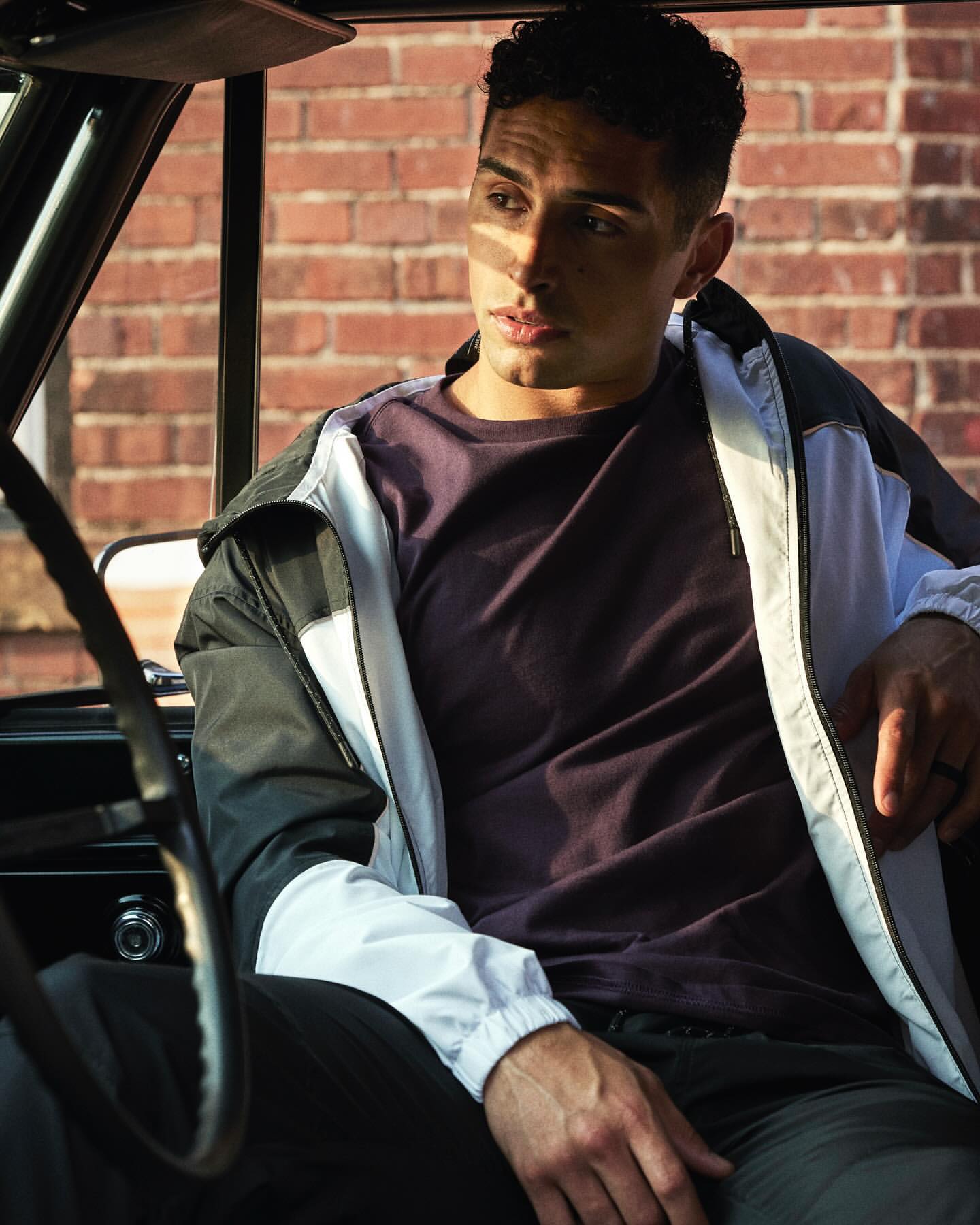 Man sits inside an old car wearing a windbreaker and a purple t-shirt, with a brick building visible in the background.