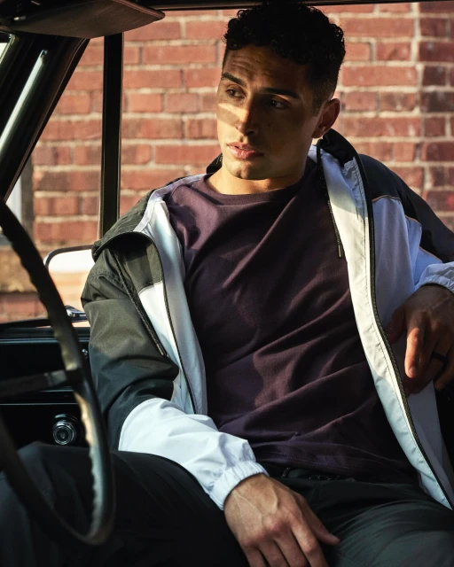 Man sits inside an old car wearing a windbreaker and a purple t-shirt, with a brick building visible in the background.