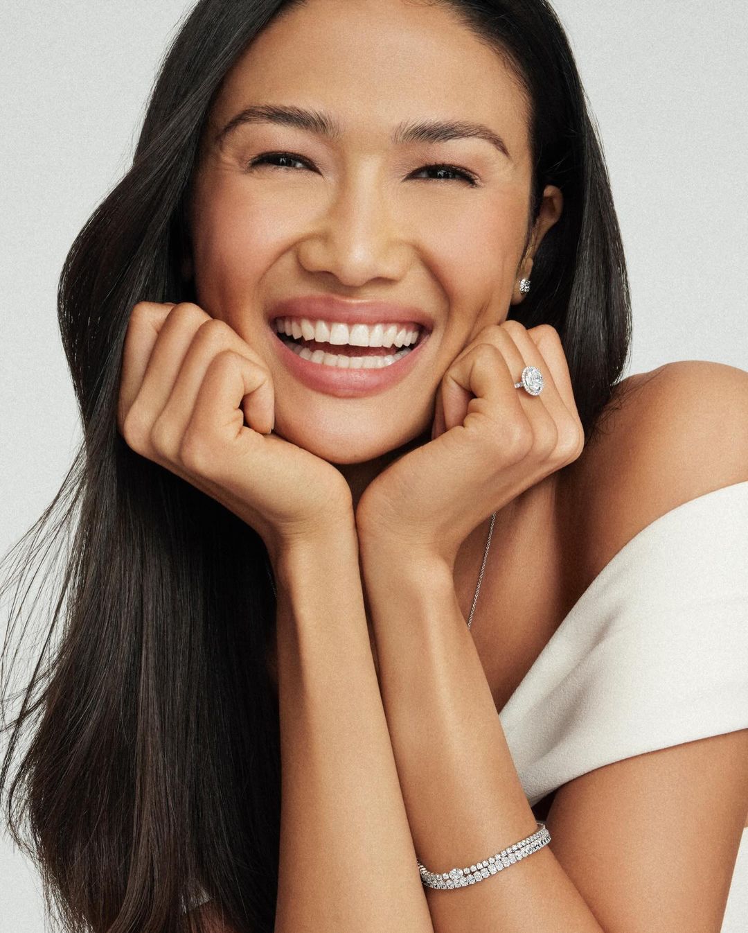 Close-up portrait of a smiling Asian woman with dark hair wearing jewelry, posing with hands near her face against a light background.