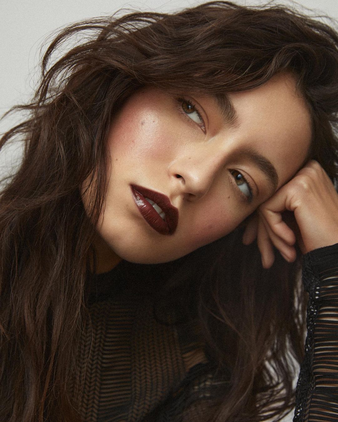 Close-up portrait of a woman with dark wavy hair, wearing a dark top. She is gazing upwards against a neutral background.