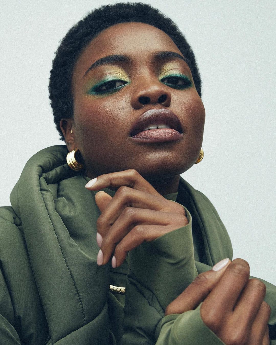 Close-up portrait of a woman with green eyeshadow and olive-green puffer jacket against a neutral background in a studio environment.