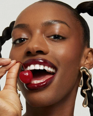 Close-up of a dark-skinned woman with braided pigtails, bright makeup, gold earrings, holding a cherry near her mouth, smiling.
