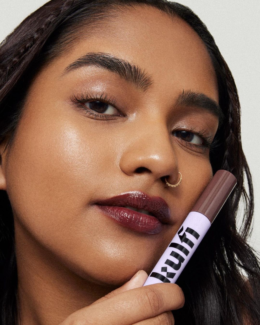 A dark-skinned woman is holding a purple and white makeup applicator in a close-up beauty product shot with glossy lips and a nose ring.