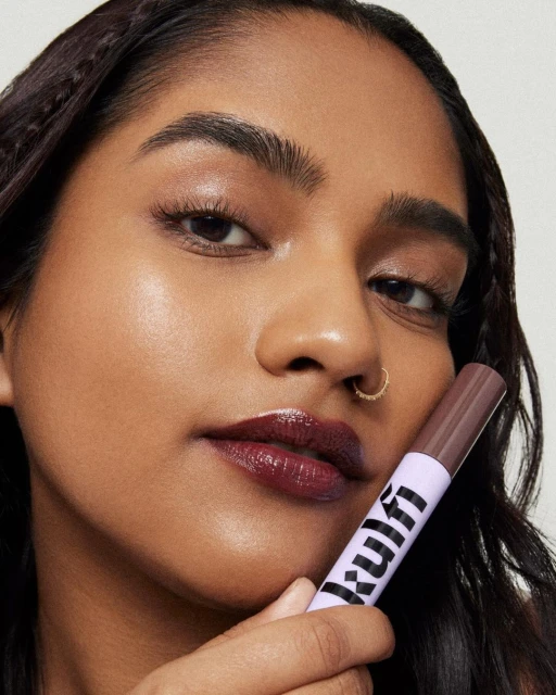 A dark-skinned woman is holding a purple and white makeup applicator in a close-up beauty product shot with glossy lips and a nose ring.