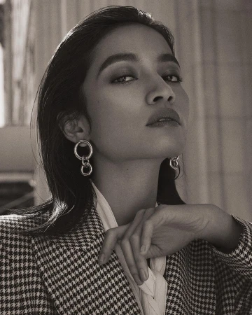 Striking portrait of an adult woman with dark hair and hoop earrings, wearing a houndstooth jacket and gazing intently at the camera.