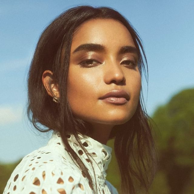 Portrait of a woman with dark hair, wearing a white lace-patterned top, looking at the camera against a blue sky and green background.