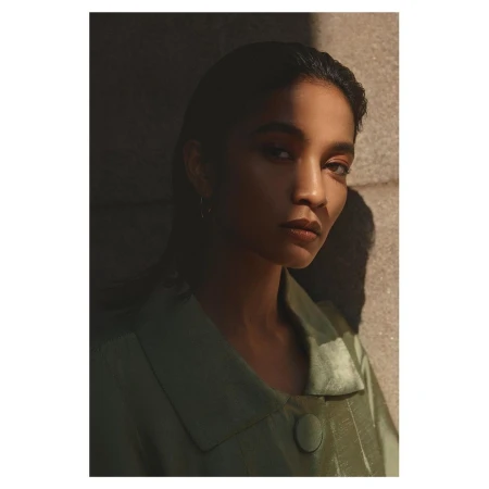 Portrait of a woman wearing a green collared jacket against a stone wall with dramatic directional sunlight.