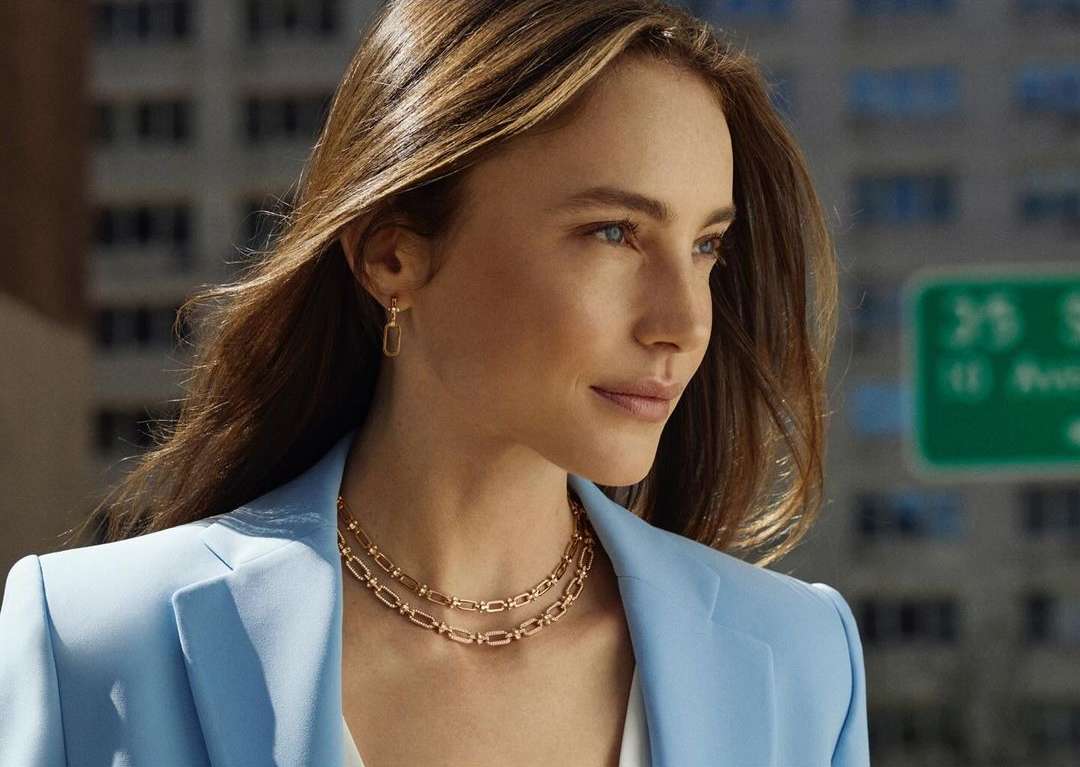 Close-up portrait of a woman wearing a light blue blazer, layered gold necklaces, and matching rectangular link earrings.