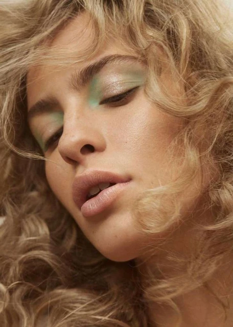 Close-up portrait of a woman with blonde curly hair, wearing light green eyeshadow and nude lipstick. Beauty shot with soft lighting.