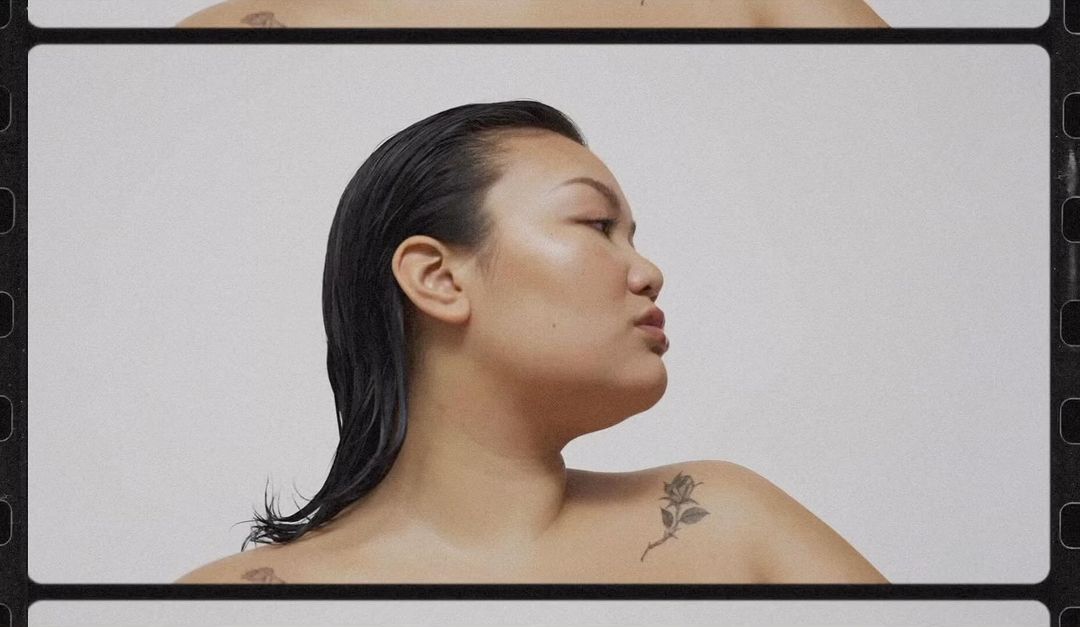 Asian woman with wet dark hair, shoulder tattoo, and tan skin posing against a plain white background in a stylized film frame.