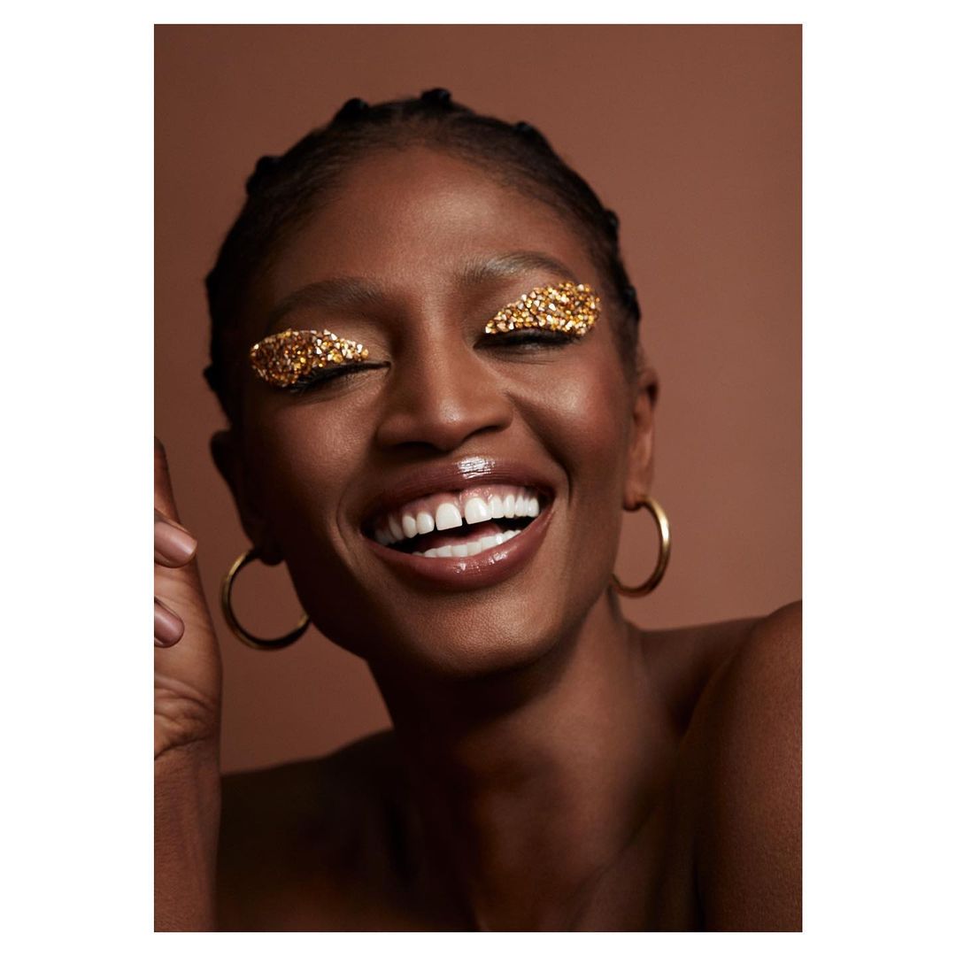 Close-up studio shot of a smiling Black woman with gold glitter eyeshadow, hoop earrings, and slicked-back hair against a brown backdrop.