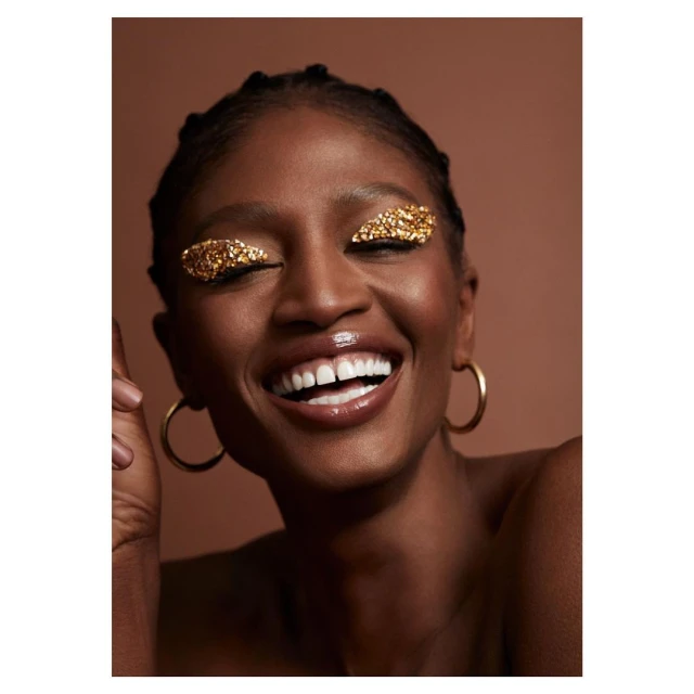 Close-up studio shot of a smiling Black woman with gold glitter eyeshadow, hoop earrings, and slicked-back hair against a brown backdrop.