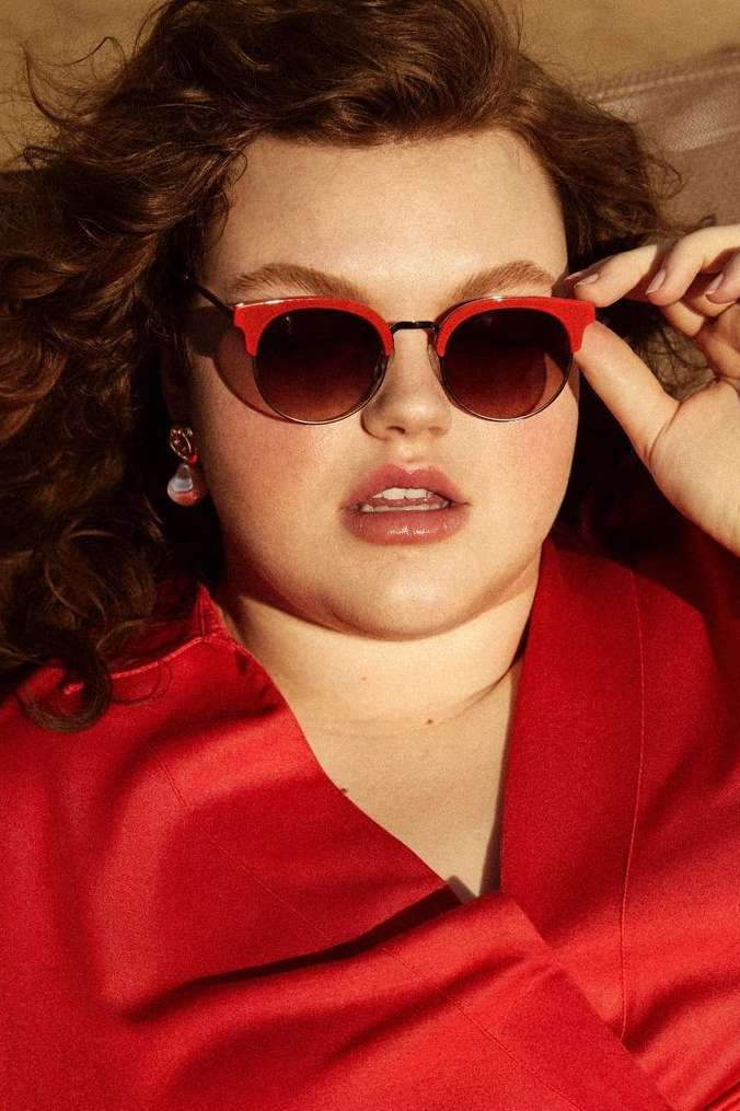 Close-up of a young woman with curly hair wearing red sunglasses and a red jacket, posing indoors. Beauty and fashion portrait.