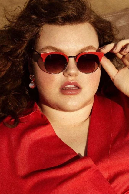 Close-up of a young woman with curly hair wearing red sunglasses and a red jacket, posing indoors. Beauty and fashion portrait.