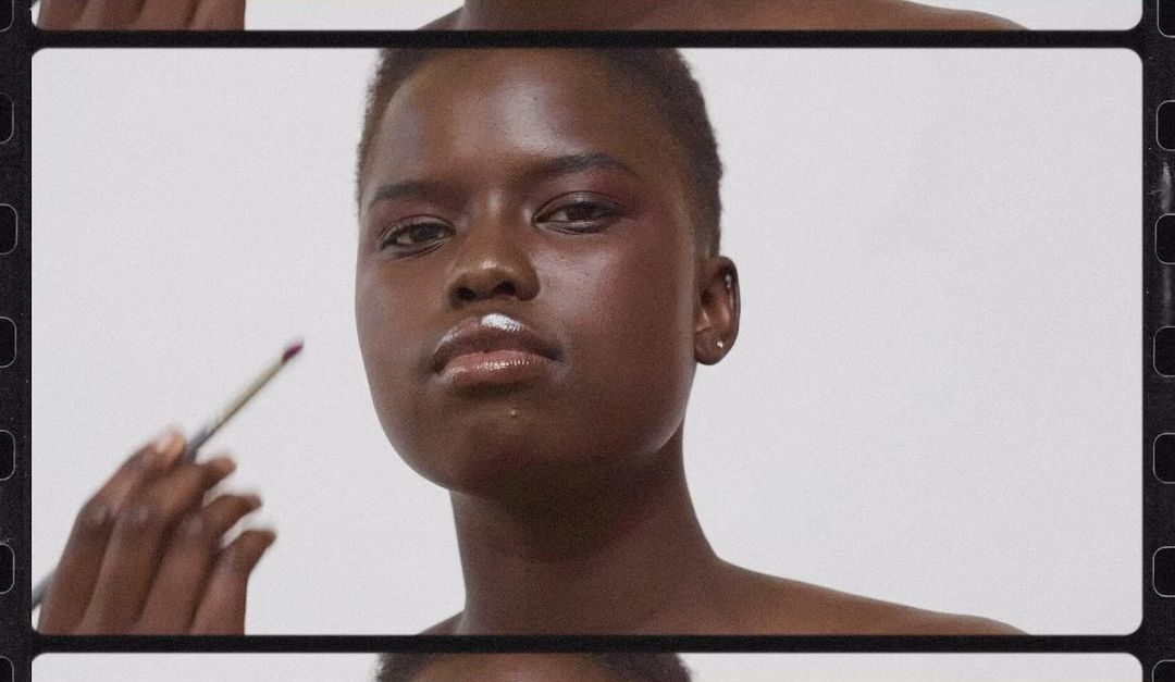Close-up of an African-descent adult female having makeup applied by a hand holding a brush in a beauty or cosmetic context.