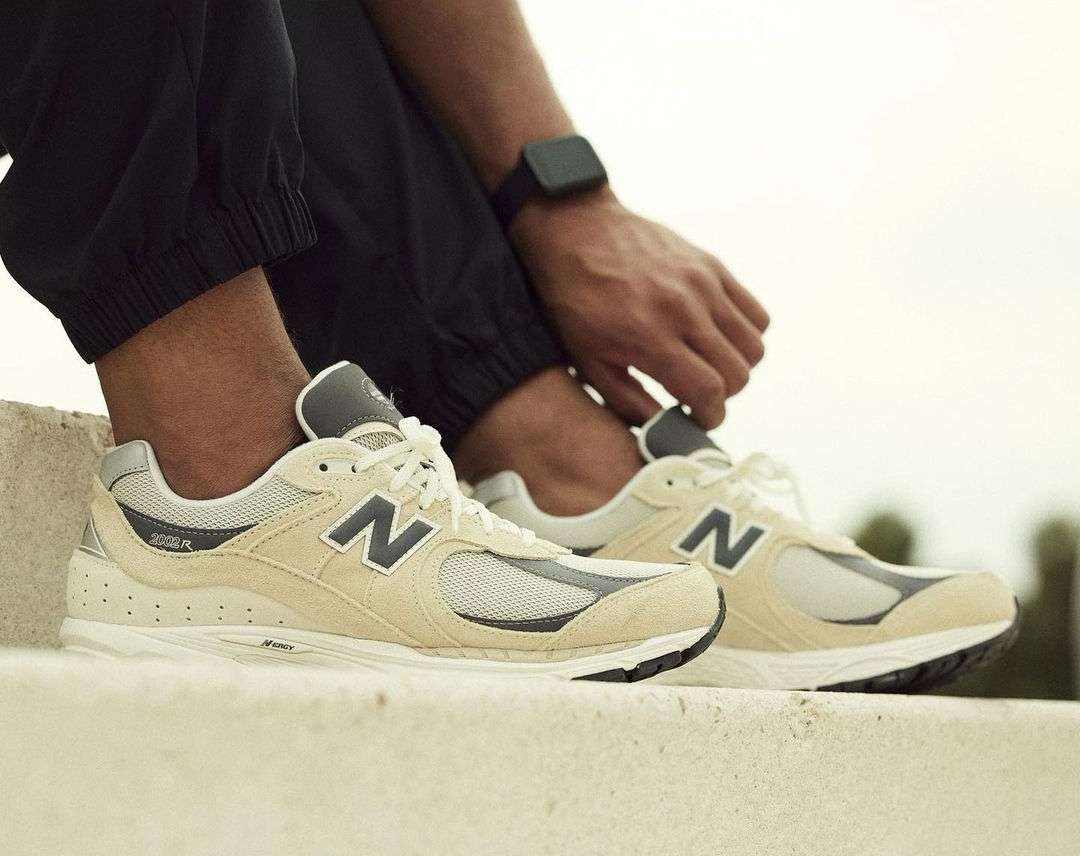 Tan and grey New Balance sneakers with a large N logo, sitting on a concrete ledge with a person tying the laces. Close-up.