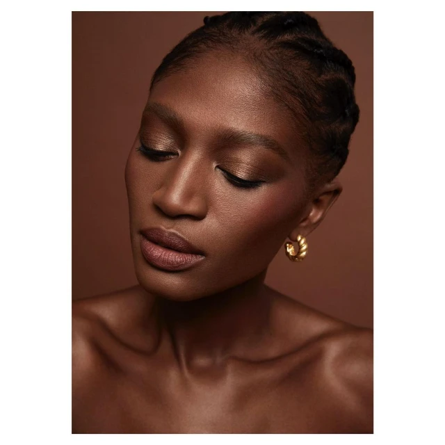 Close-up beauty portrait of a woman with dark skin, gold hoop earrings, and braided hair against a warm brown studio backdrop.