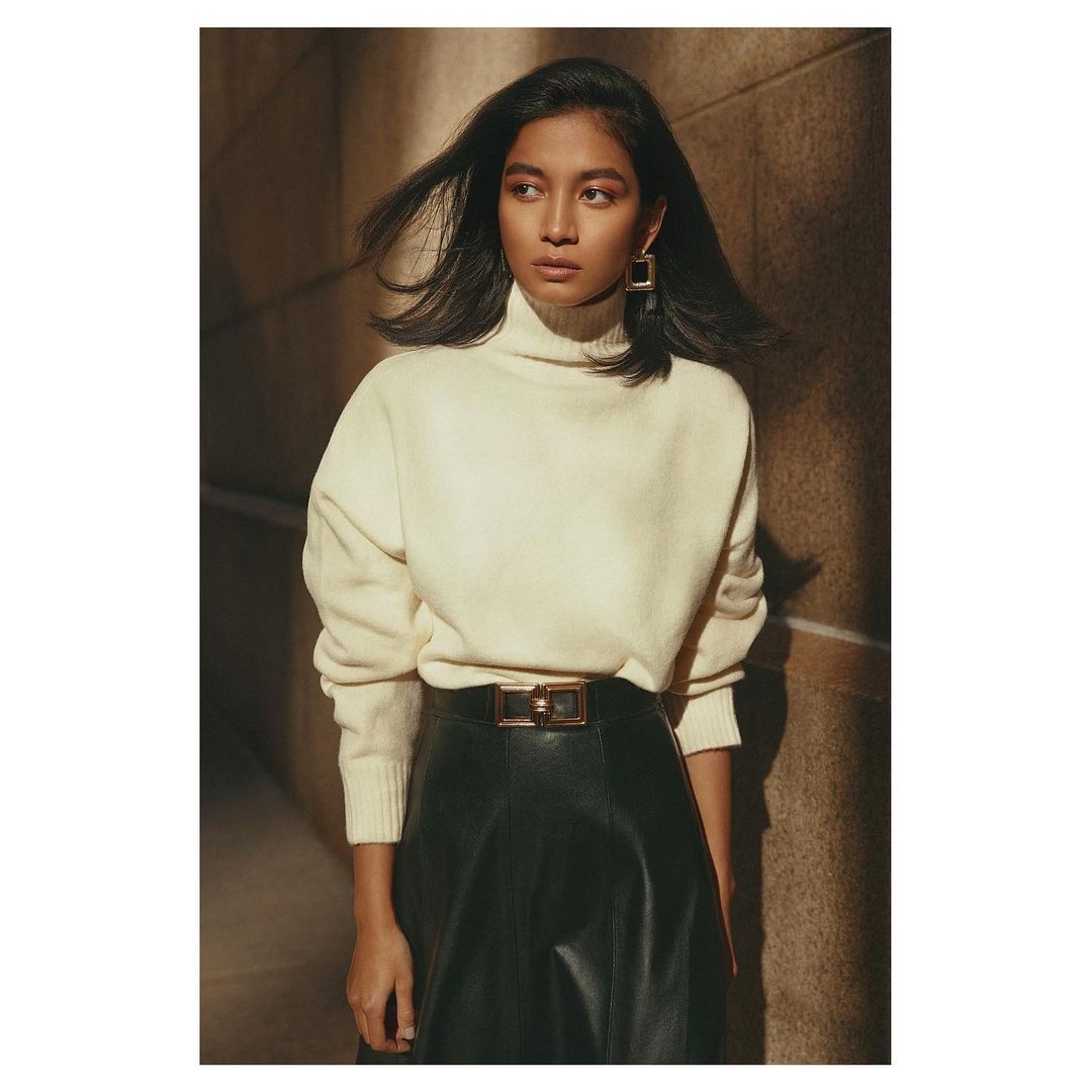 Portrait of a young woman with long hair wearing a turtleneck sweater, leather skirt with belt, and gold earrings, posing in a warm-toned indoor space.