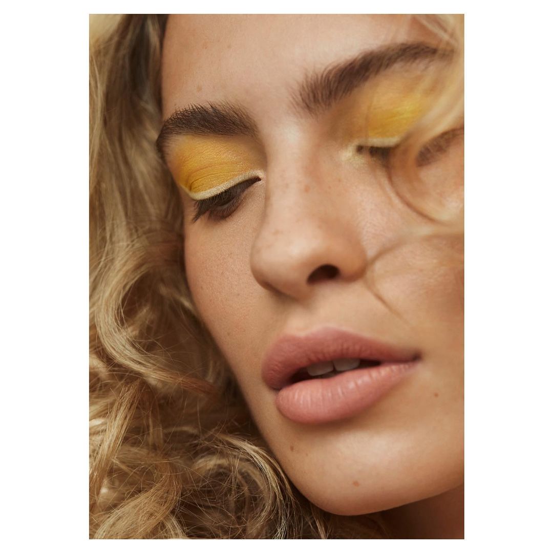 Close-up portrait of a woman with blonde curly hair and yellow eyeshadow, focusing on beauty and cosmetic elements in a studio setting.