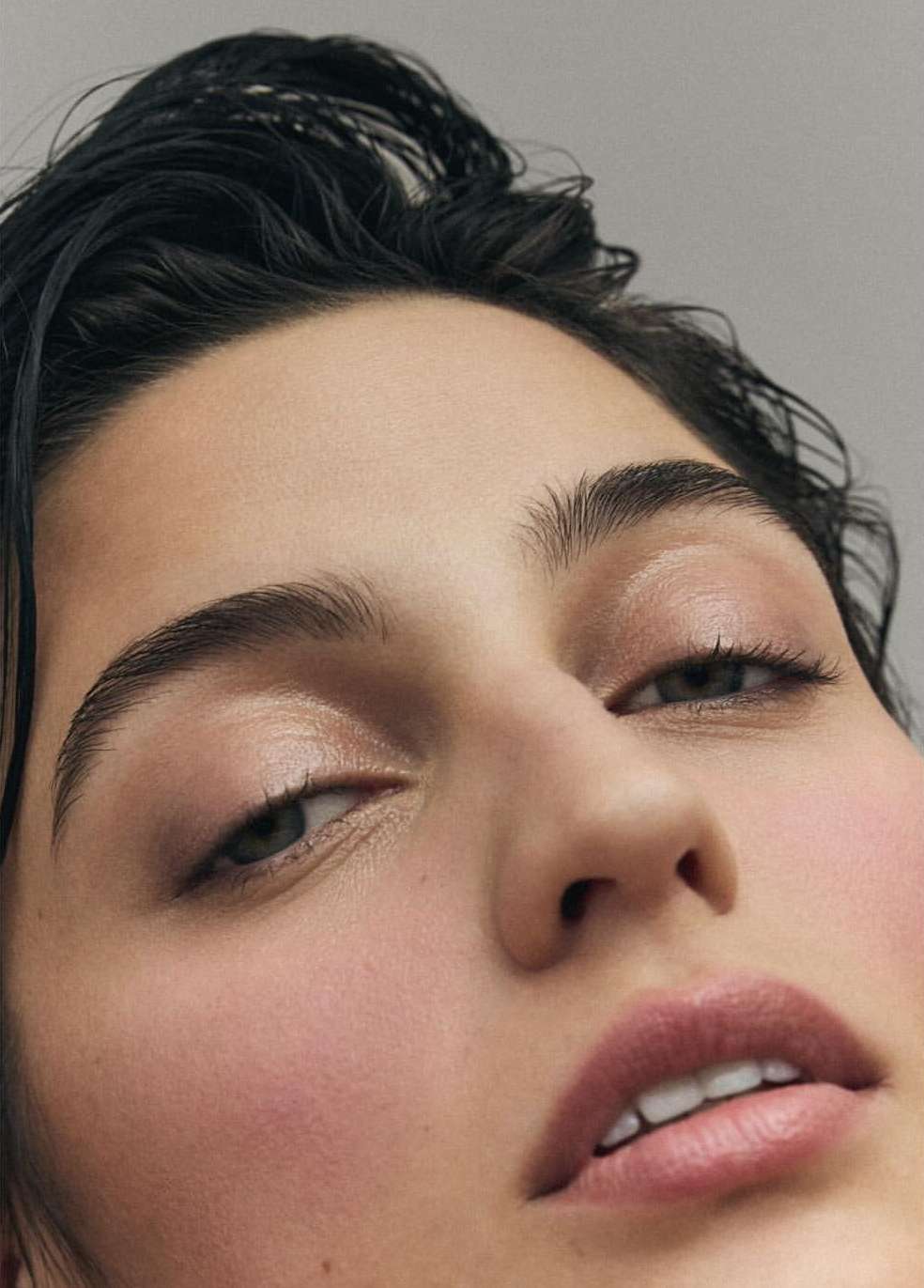 Close-up portrait of a young woman with dark hair, fair skin, and natural makeup, gazing upwards against a neutral grey background.