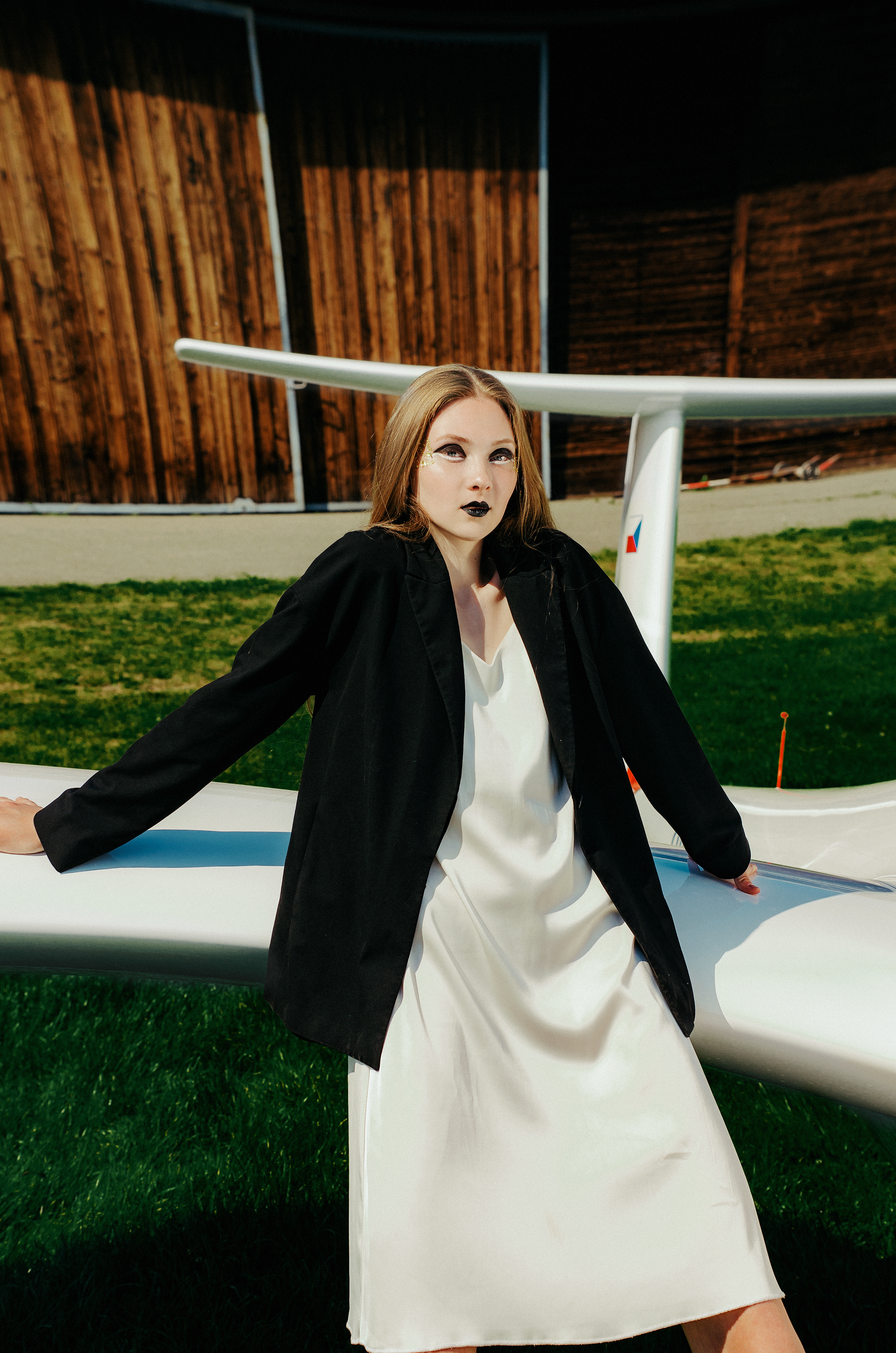 Woman in white slip dress and black blazer posing on airplane wing outdoors