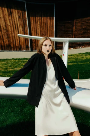 Woman in white slip dress and black blazer posing on airplane wing outdoors
