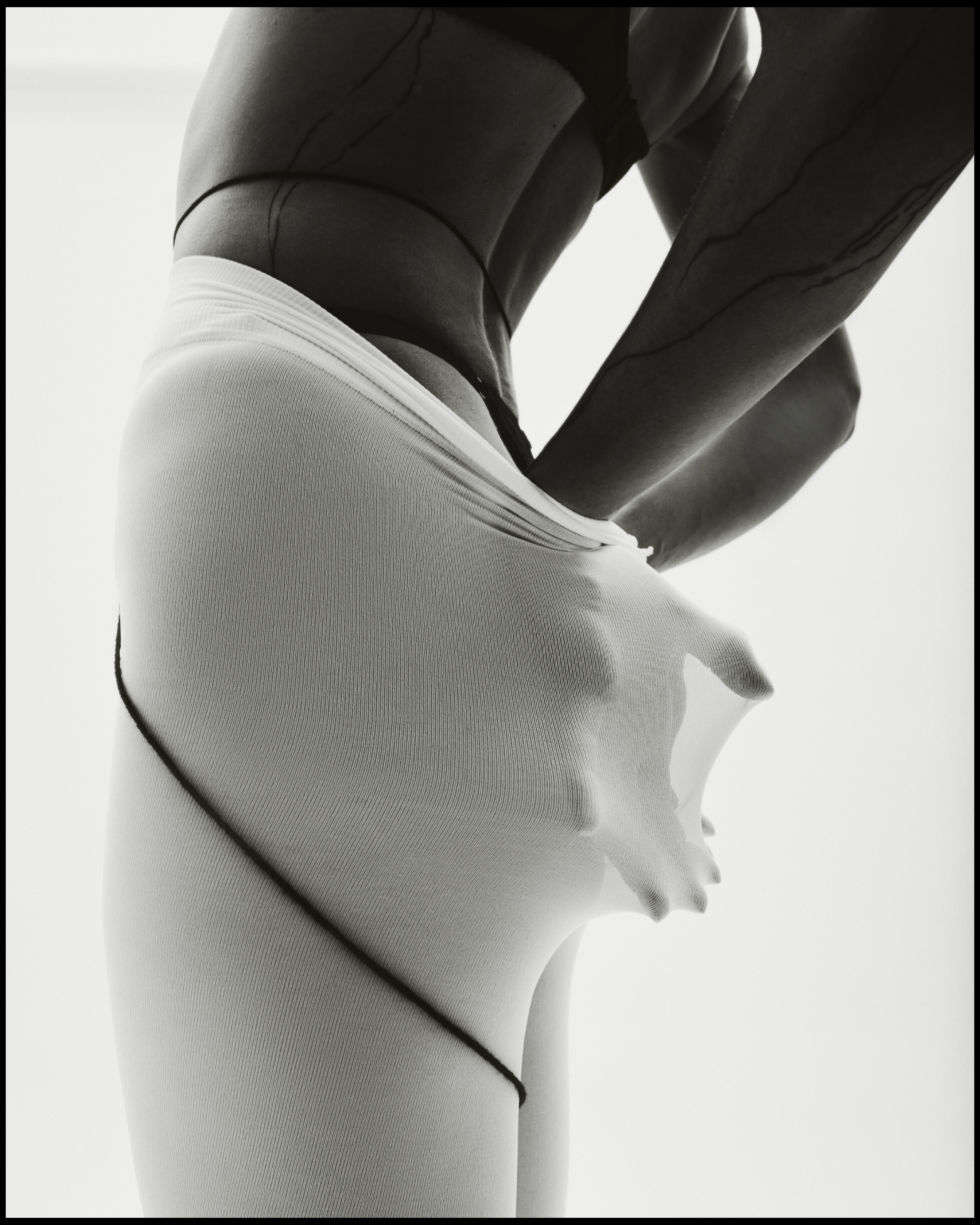 Black and white shot of a person wearing sheer white tights, stretching the fabric outwards with their hand against a white backdrop.