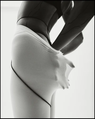 Black and white shot of a person wearing sheer white tights, stretching the fabric outwards with their hand against a white backdrop.