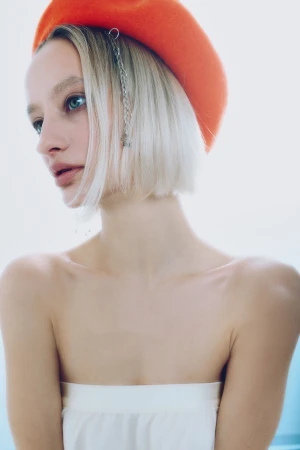 Blonde woman with a short bob haircut wearing a vibrant orange beret and white strapless top looking to the side.