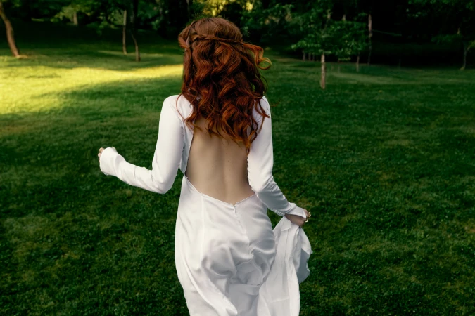 Rear view of a woman with long red hair wearing a backless white dress running across a grassy park.
