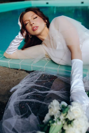Woman in a white sheer gown and white satin gloves reclining by the tiled edge of a turquoise pool with a floral bouquet.