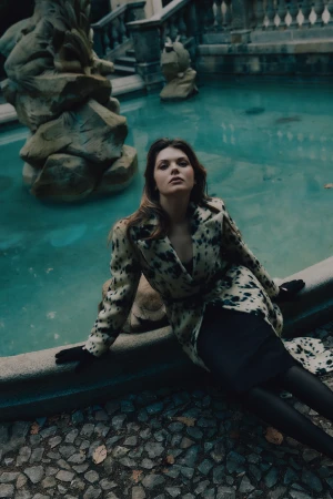 Woman in a spotted print coat and black gloves reclining against an ornate stone fountain basin filled with teal water.