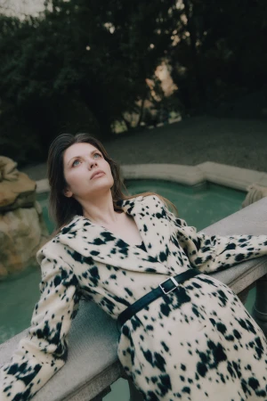 Woman in a cow-print coat leaning against a stone railing outdoors in a park.
