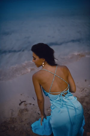 A woman in an open-back light blue satin slip dress kneels on a sandy beach shoreline near gentle ocean waves at dusk.