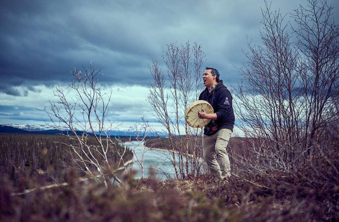 Indigenous man plays a round drum on a hill, singing with snowy mountains, trees, and dramatic sky in the background on an overcast day.