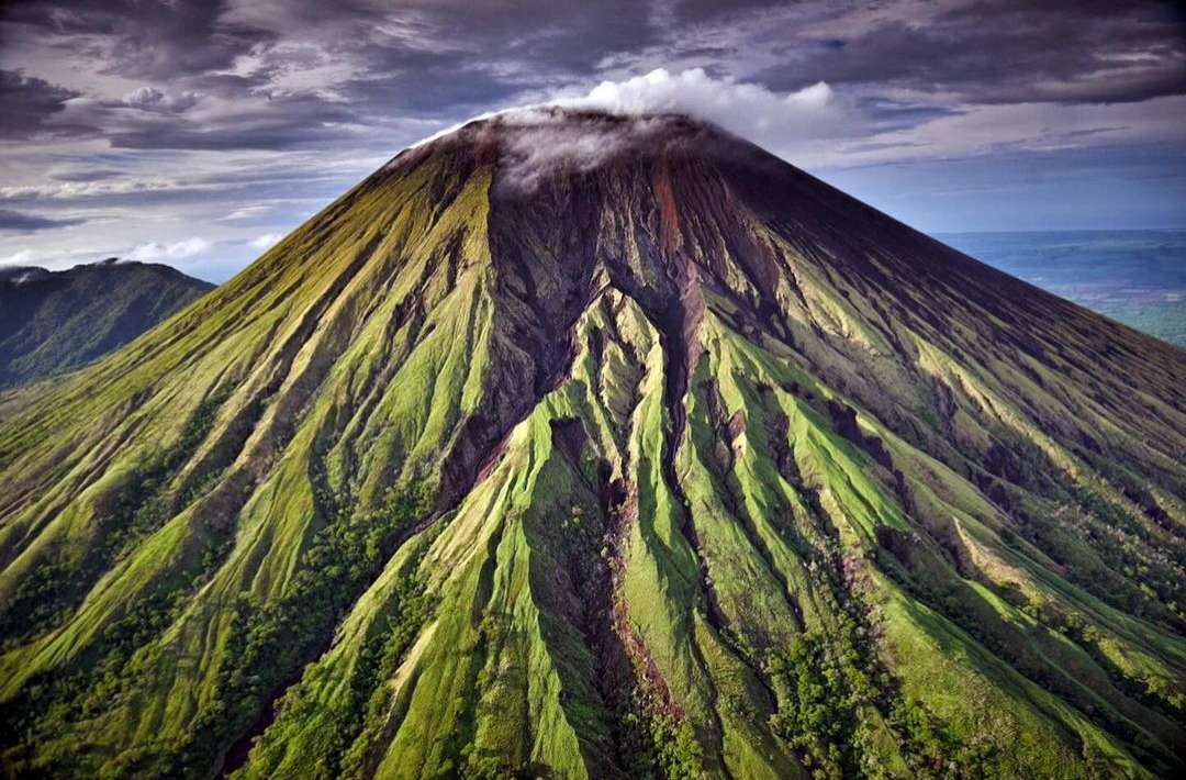 Dramatic aerial view of a lush green mountain with a cloudy peak. The landscape is dominated by the mountain and its vibrant colors.