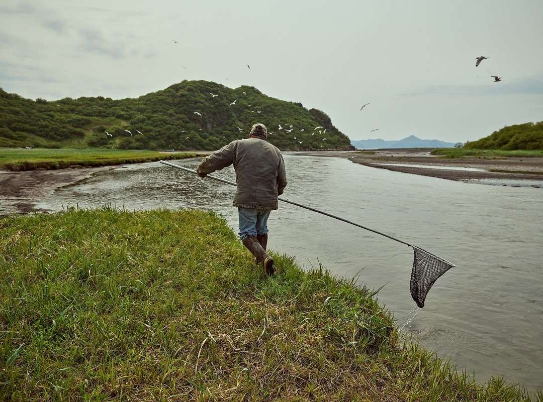 A man in a jacket and boots stands at the edge of a river with a fishing net, with grassy banks, a forested hill, and birds in the sky.