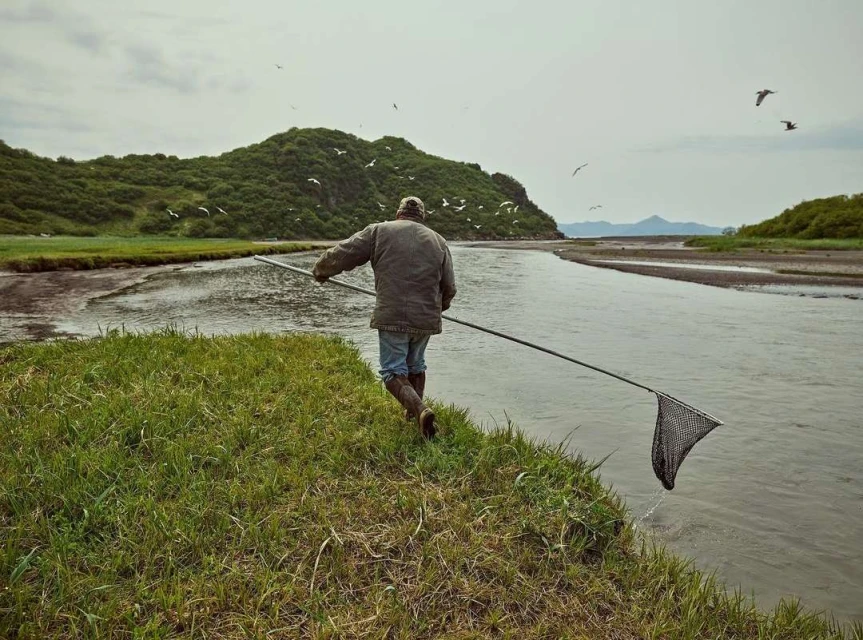 A man in a jacket and boots stands at the edge of a river with a fishing net, with grassy banks, a forested hill, and birds in the sky.