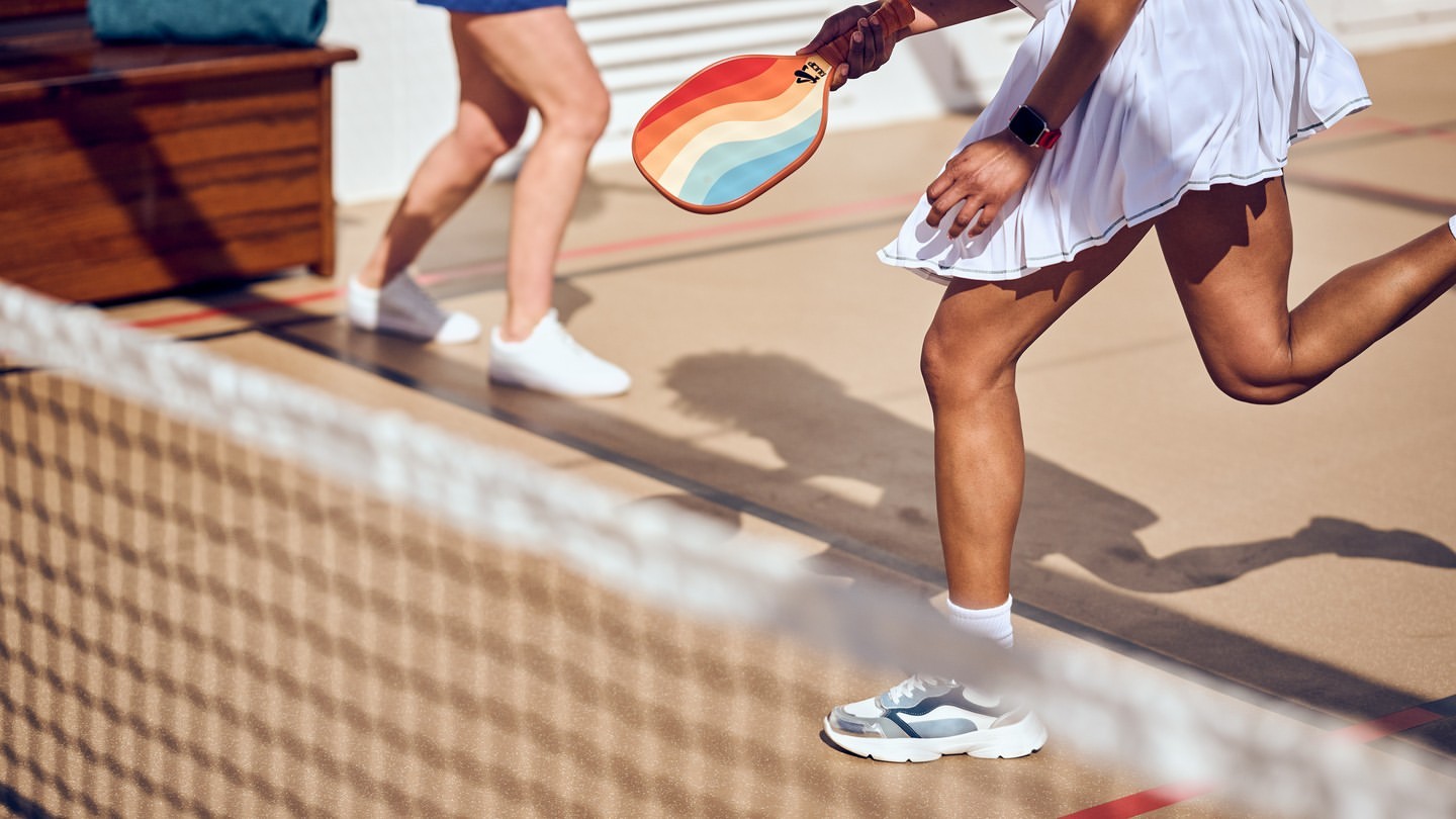 Two women play paddleball on a sand-colored court. One woman holds a rainbow paddle, and the other woman runs in the background.