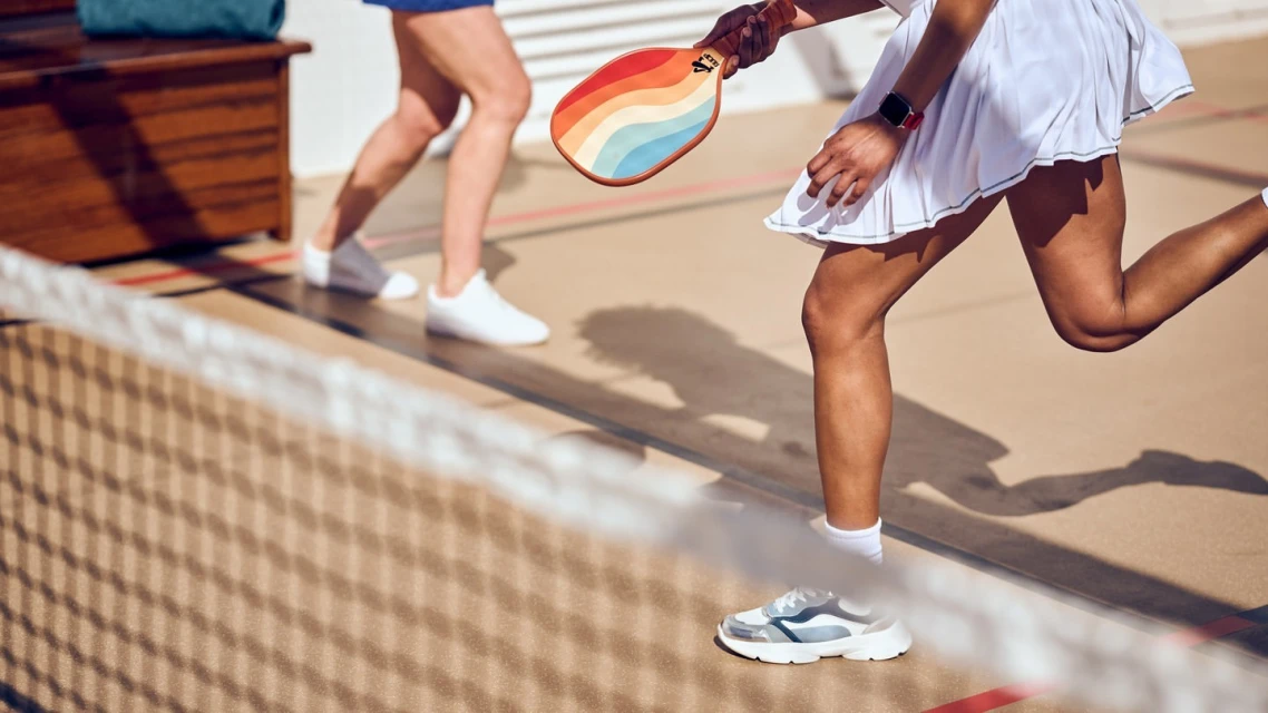 Two women play paddleball on a sand-colored court. One woman holds a rainbow paddle, and the other woman runs in the background.