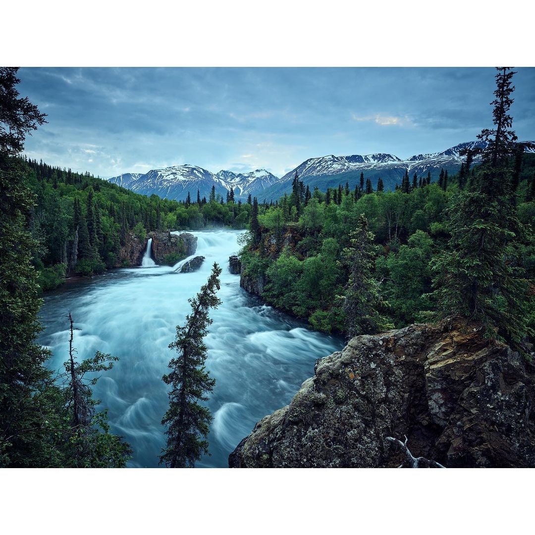 Scenic view of a river cascading through a forest landscape, with snow-capped mountains visible in the background under a cloudy sky.