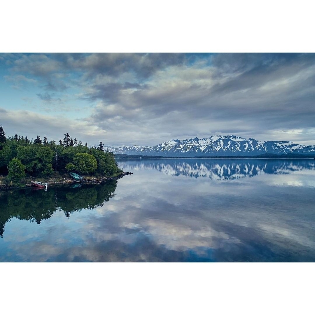 Scenic view of a calm lake reflecting the sky, with lush green trees on the shoreline and snow-capped mountains in the distance.